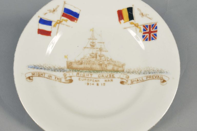 2015.9.1-i White Grosvenor china World War One Commemorative Plate with a gold rim, made in late 1914. On it are flags of the Allies (France, Russia, Belgium and Britain), a ship and the words: ‘Might in the right cause shall prevail. European War 1914 & 15’. The maker is Jackson & Gosling, England.