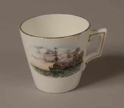 Demi Tasse white porcelain coffee cup with a gold rim around the top. Front of cup is decorated with a picture of a World War One battleship firing a broadside with painted words: ‘H.M Battleship in action’.