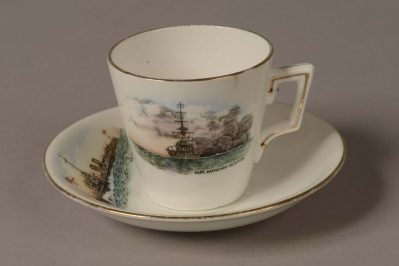 GEI-0023_GEI-0022-e1422843434422 Demi Tasse white porcelain coffee cup and saucer with a gold rim around the top. Front of cup is decorated with a picture of a World War One battleship firing a broadside with painted words: ‘H.M Battleship in action’. Top of saucer is decorated with a picture of a World War One torpedo boat with a white ensign at the jack. Words on the saucer read: ‘H.M Torpedo Boat’.