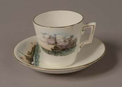 Demi Tasse white porcelain coffee cup and saucer with a gold rim around the top. Front of cup is decorated with a picture of a World War One battleship firing a broadside with painted words: ‘H.M Battleship in action’. Top of saucer is decorated with a picture of a World War One torpedo boat with a white ensign at the jack. Words on the saucer read: ‘H.M Torpedo Boat’.